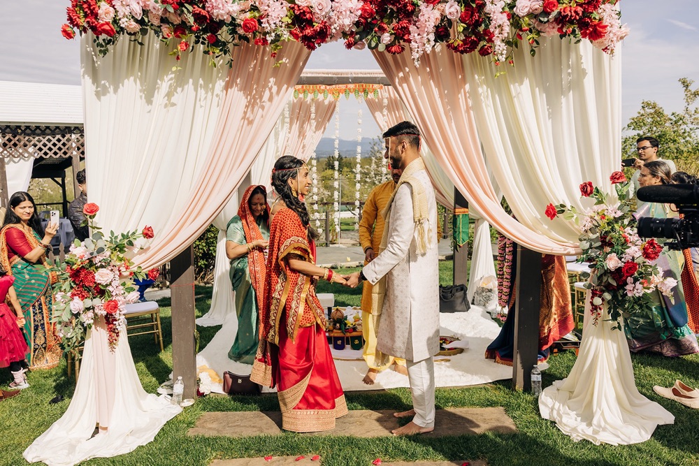 Bride and groom facing each other under a blush and ivory Mandap with red and pink silk florals at Eagle Mountain Venue