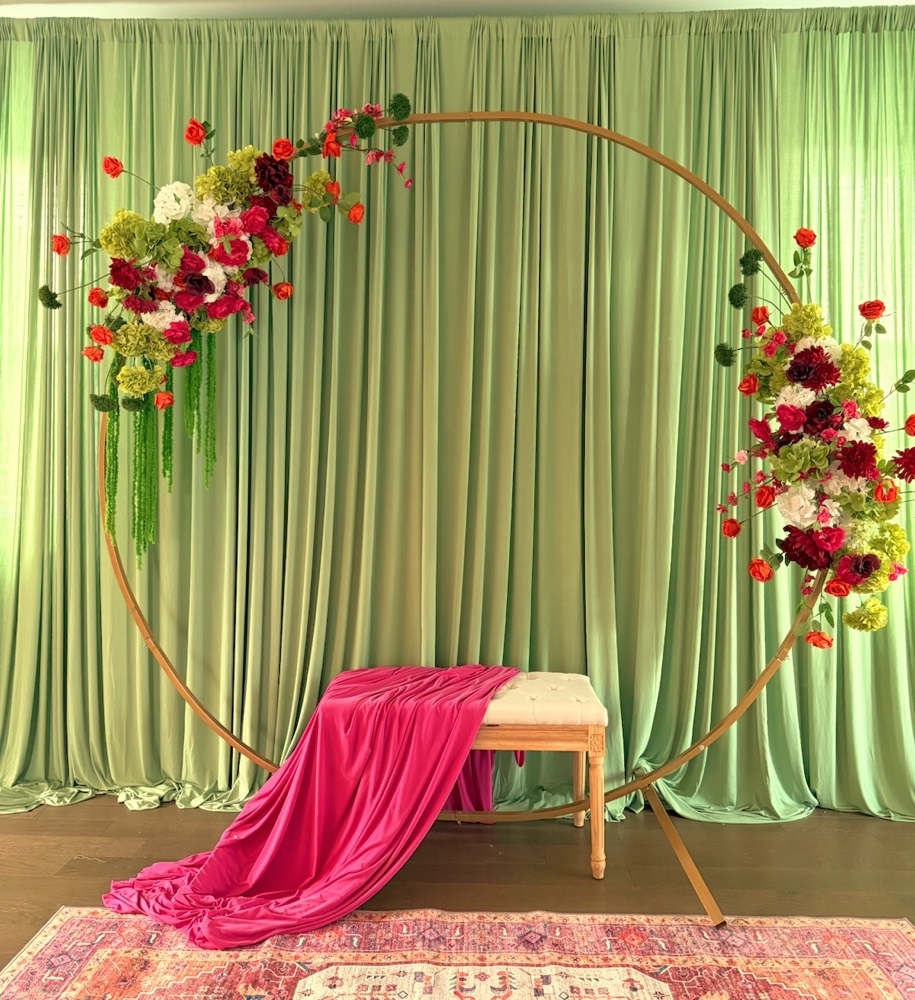 Mehndi backdrop with sage green drapery, a gold ring arch with red and pink silk florals and a bench draped in fuchsia