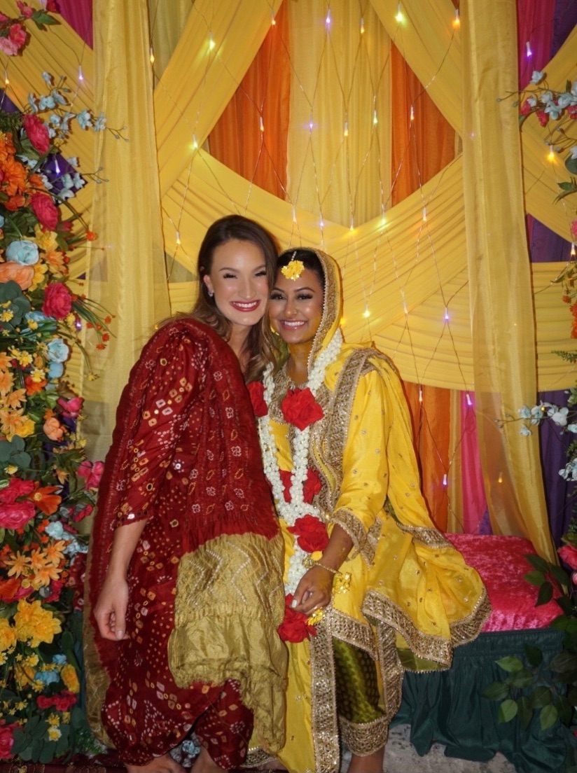 A bride in yellow with floral jewelry and a guest at her mehndi, in front of yellow, orange and purple drapery with string lights and silk florals