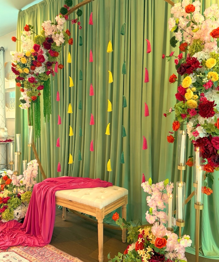 Mehndi decor with sage green drapery, colored tassels, red, pink and yellow silk florals, candle stands and a tufted bench draped in fuchsia